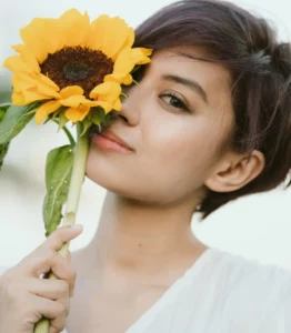 Charming woman with short brown hair and light makeup holding fresh sunflower and looking at camera